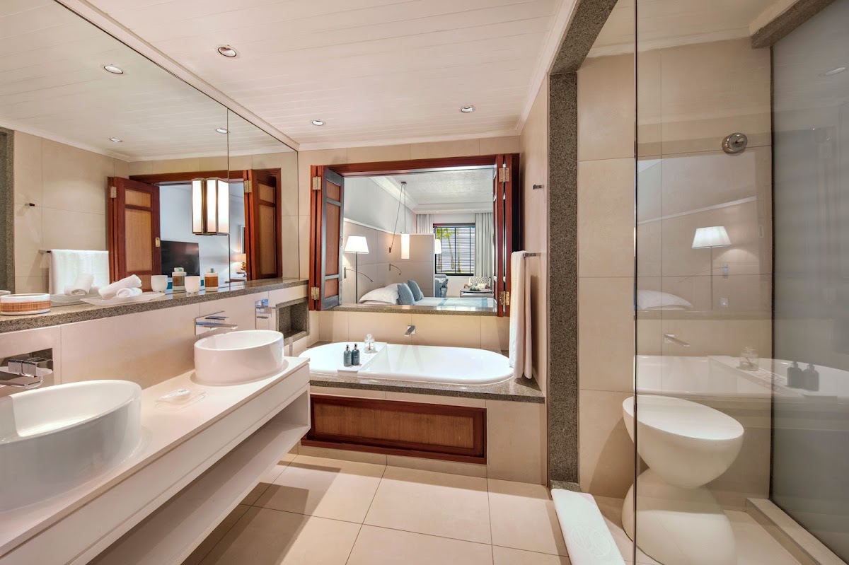 Bathroom with dual vanities, soaking tub with wood paneling, walk-in shower, and view into bedroom with wooden doors.