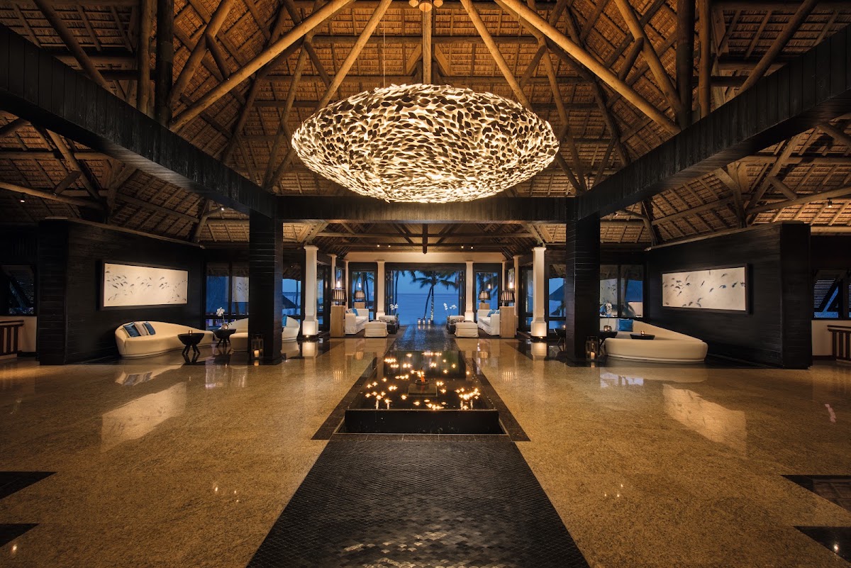 Grand lobby with woven pendant light, central fire pit, polished stone floor, and ocean views through open colonnade at dusk.