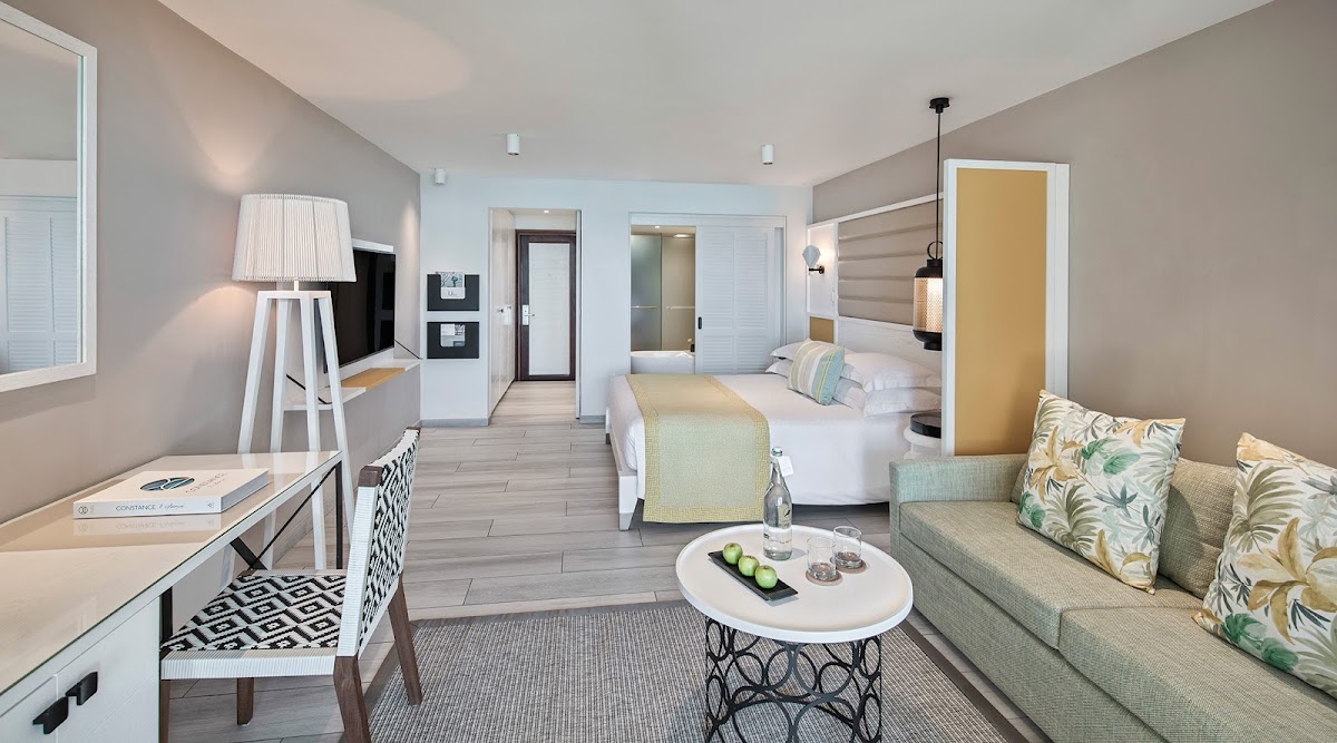 Spacious resort suite with two beds, seating area with tropical-patterned cushions, desk with lamp, and modern bathroom entrance.