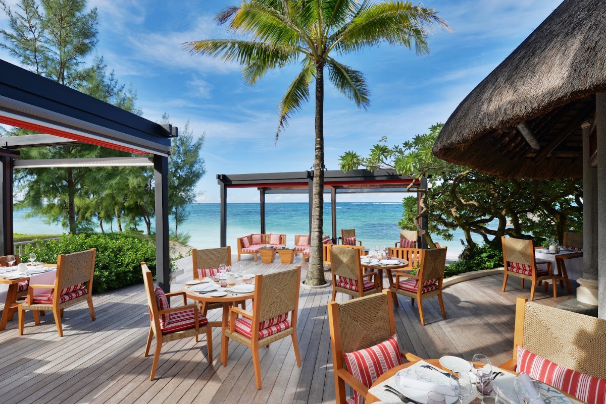Beachfront dining pavilion at Constance Belle Mare Plage with wooden chairs, tables, and ocean view beyond palm trees and thatched structures.