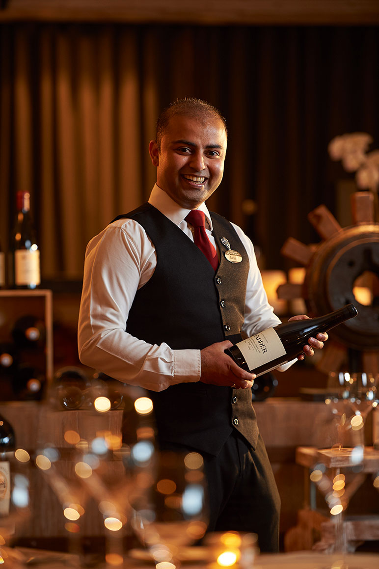 Smiling server in formal vest and tie holds a wine bottle at a candlelit dining table.