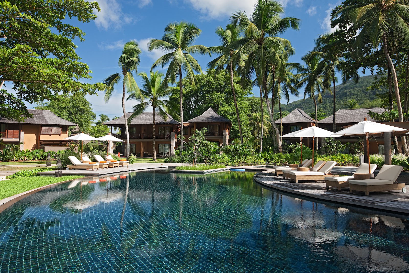 Long pool with blue tile facing wooden bungalows, palm trees, and white umbrellas shading loungers at Constance Ephelia.