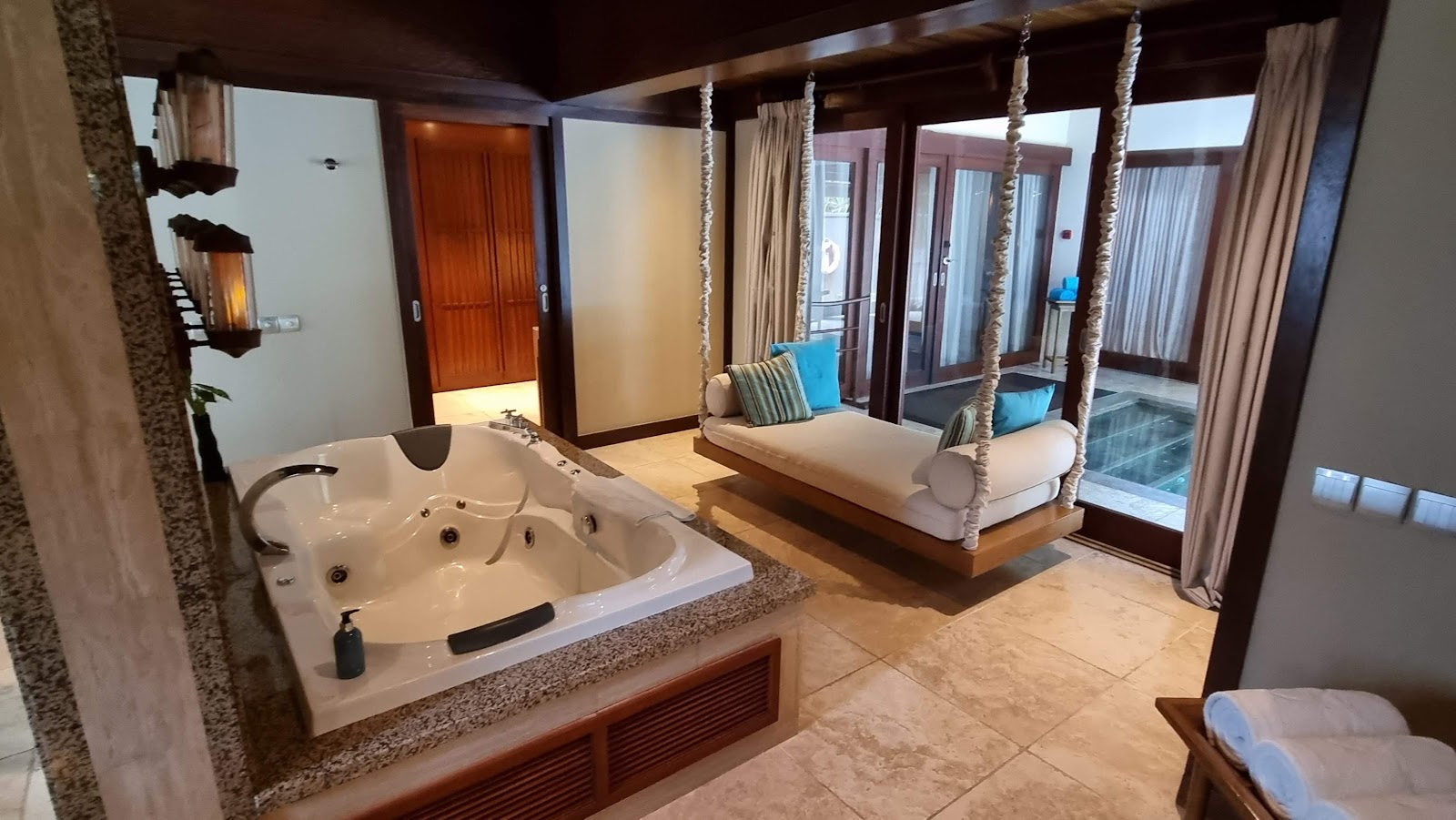 Spacious overwater bungalow bedroom at Constance Ephelia with sunken soaking tub, rope-swing bed, and ocean views through glass doors.