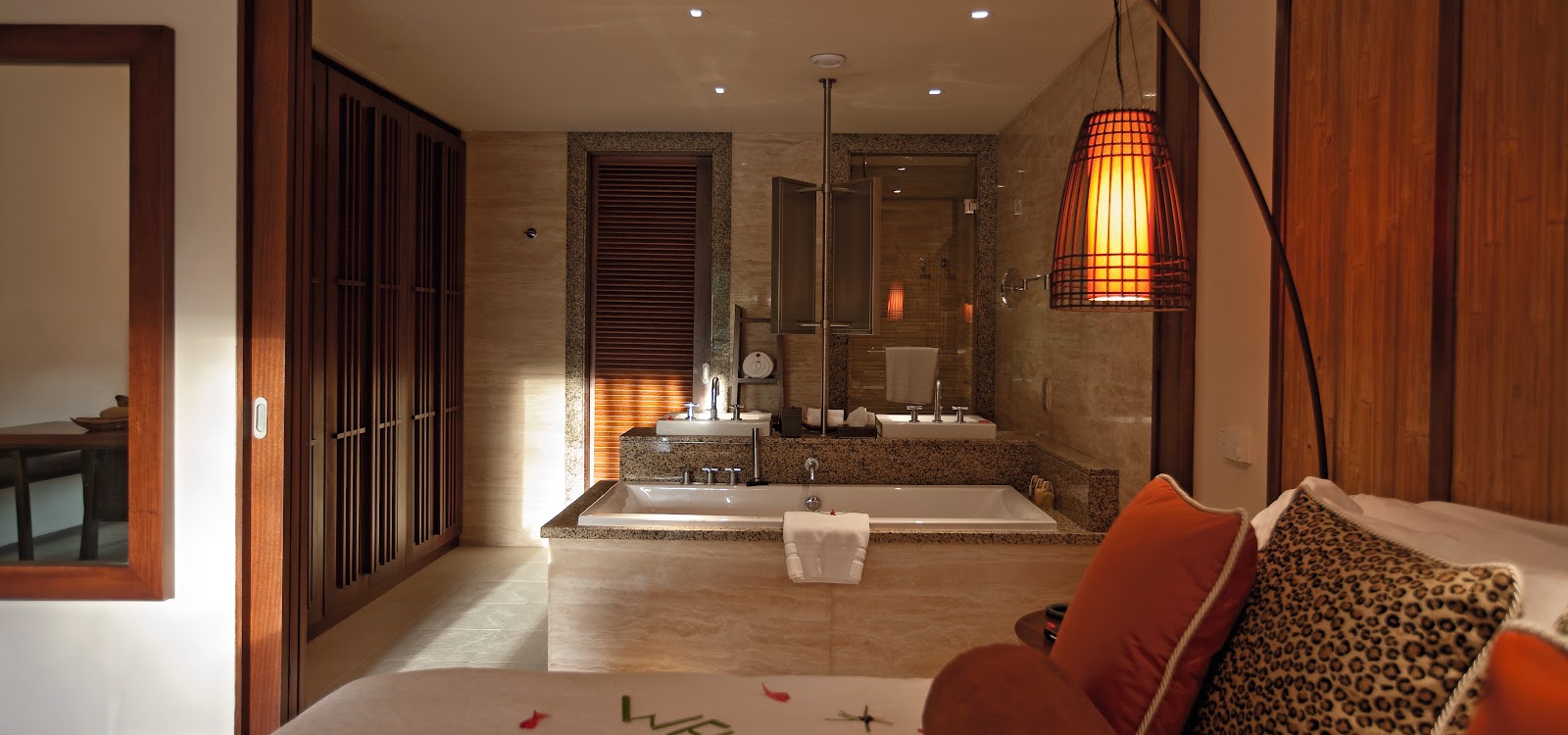 Resort villa bedroom with wooden ceiling beams, white bed with gold pillows, deck overlooking forested hillside, and en-suite bathroom with double vanity.