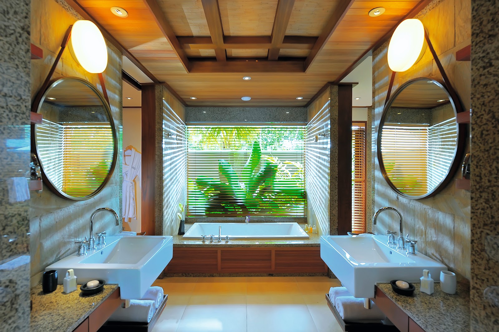 Spacious resort suite bathroom with sunken tub, dual vanity, walk-in shower, wooden accents, and hanging pendant light beside a bed with orange pillows.