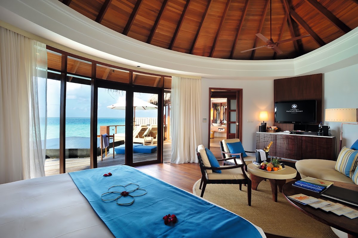 Overwater villa interior at Constance Halaveli with bed, lounge area, and glass doors opening to wooden deck overlooking turquoise ocean.