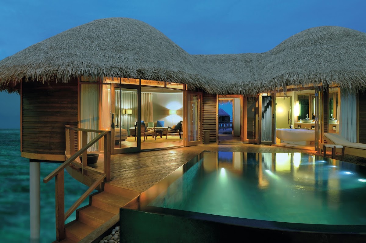 Overwater bungalows with thatched roofs and lit interiors at dusk, connected by wooden decks above turquoise lagoon water.