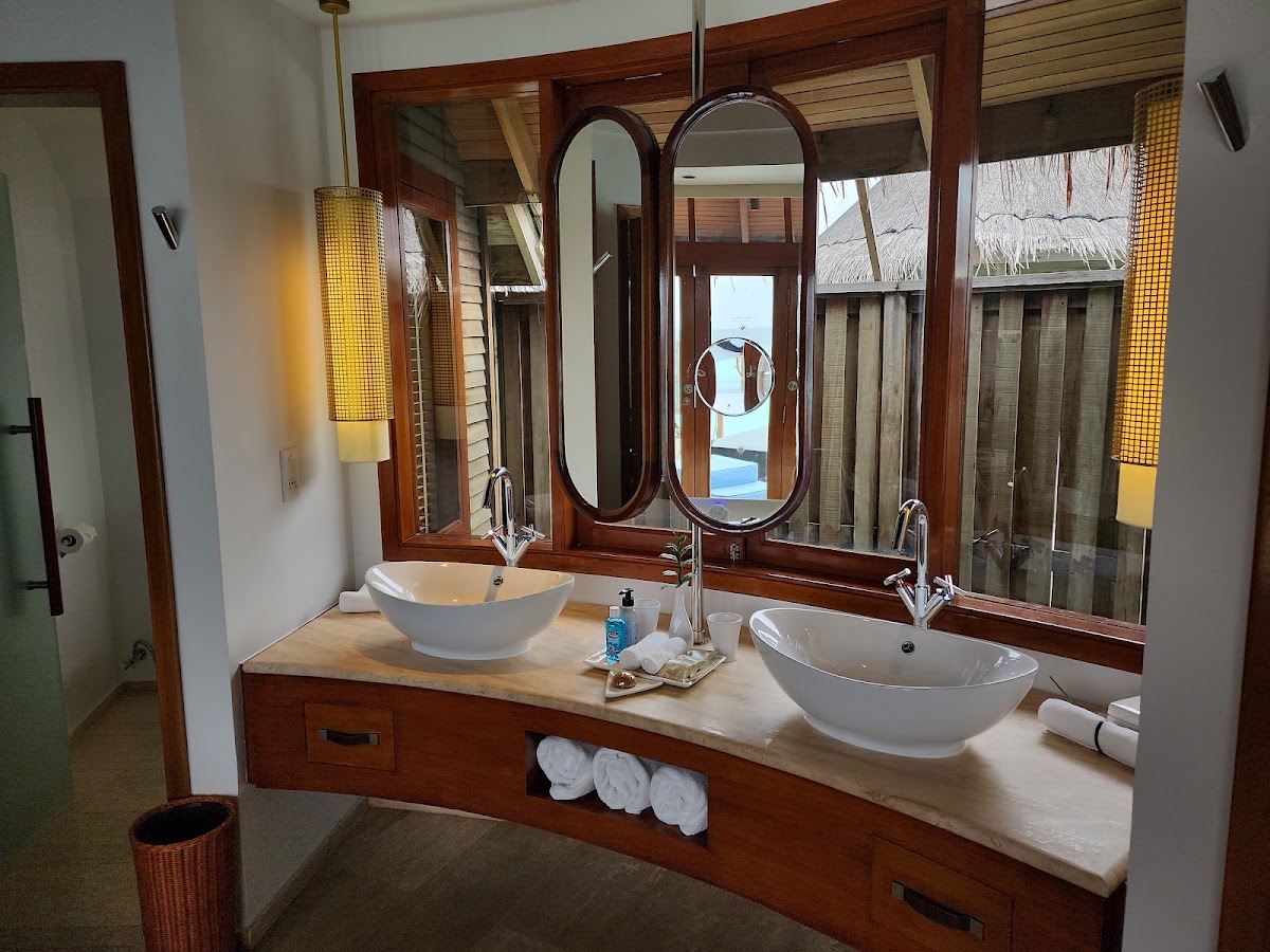Bathroom with two white vessel sinks on a wooden vanity, dual oval mirrors, and ocean view through windows with thatched roof visible outside.