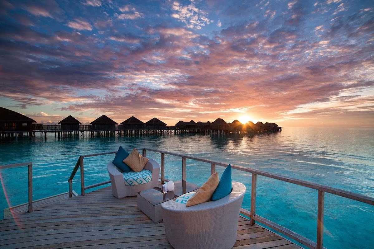 Woven lounge chairs on a wooden deck at Constance Halaveli overlook turquoise lagoon water and overwater bungalows at sunset.