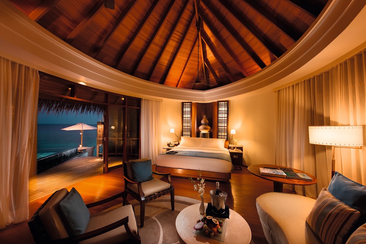 Spacious overwater villa interior at Constance Halaveli with wooden ceiling, bed, seating area, and ocean view through open doors at dusk.