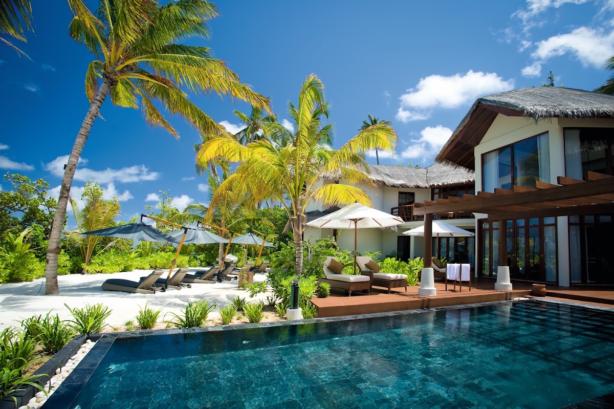 Beachfront villa at Constance Halaveli with private pool, thatched-roof bungalow, lounge chairs, white umbrellas, and palm trees under blue sky.