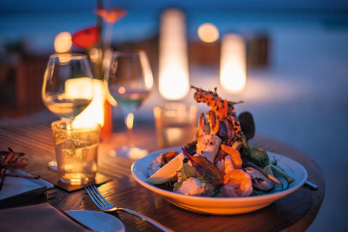 Seaside dinner table at dusk with seafood plate, wine glasses, and glowing lanterns against twilight sky.