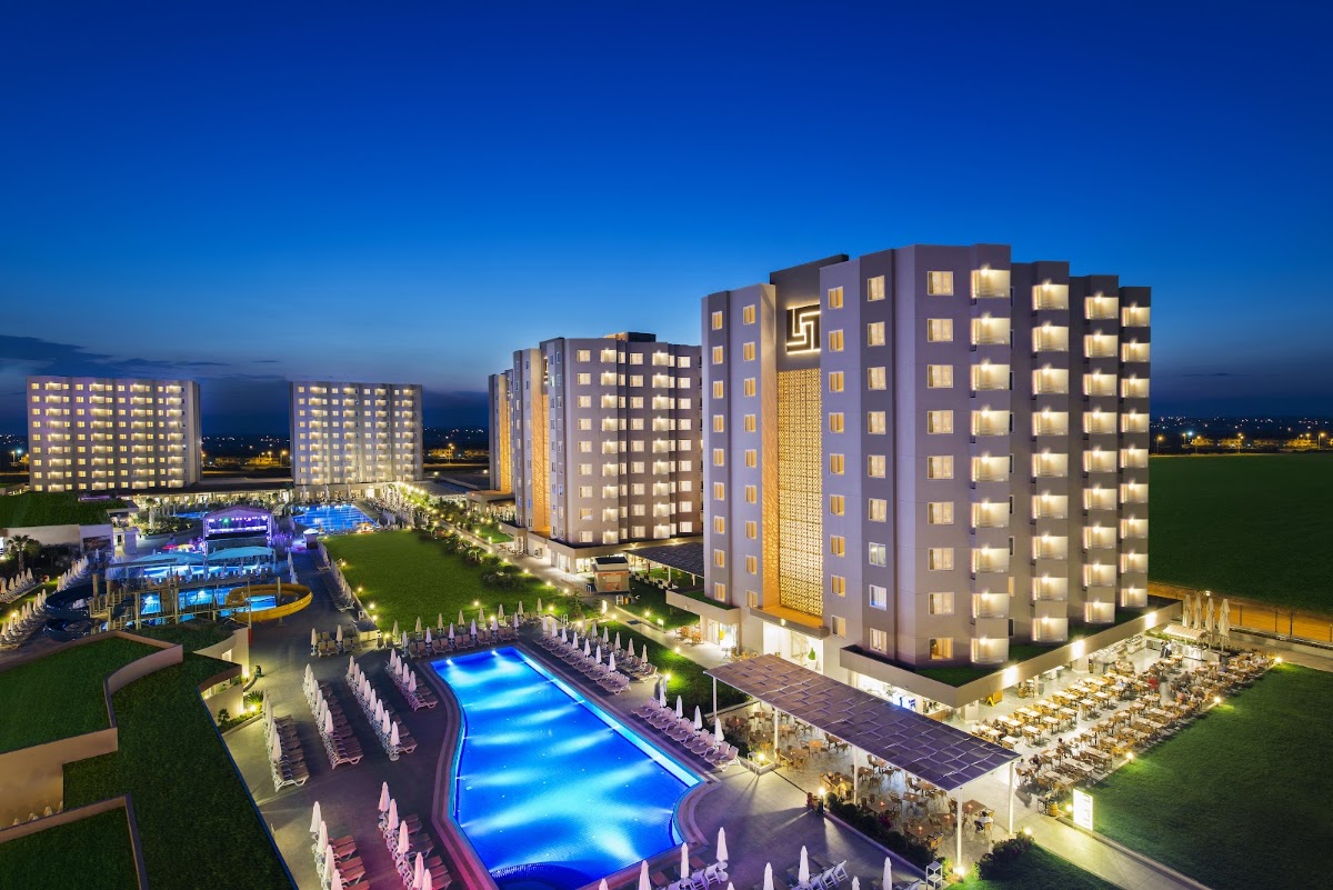 Illuminated resort buildings at dusk with glowing pools, loungers, and manicured lawns at Corendon Grand Park Lara.