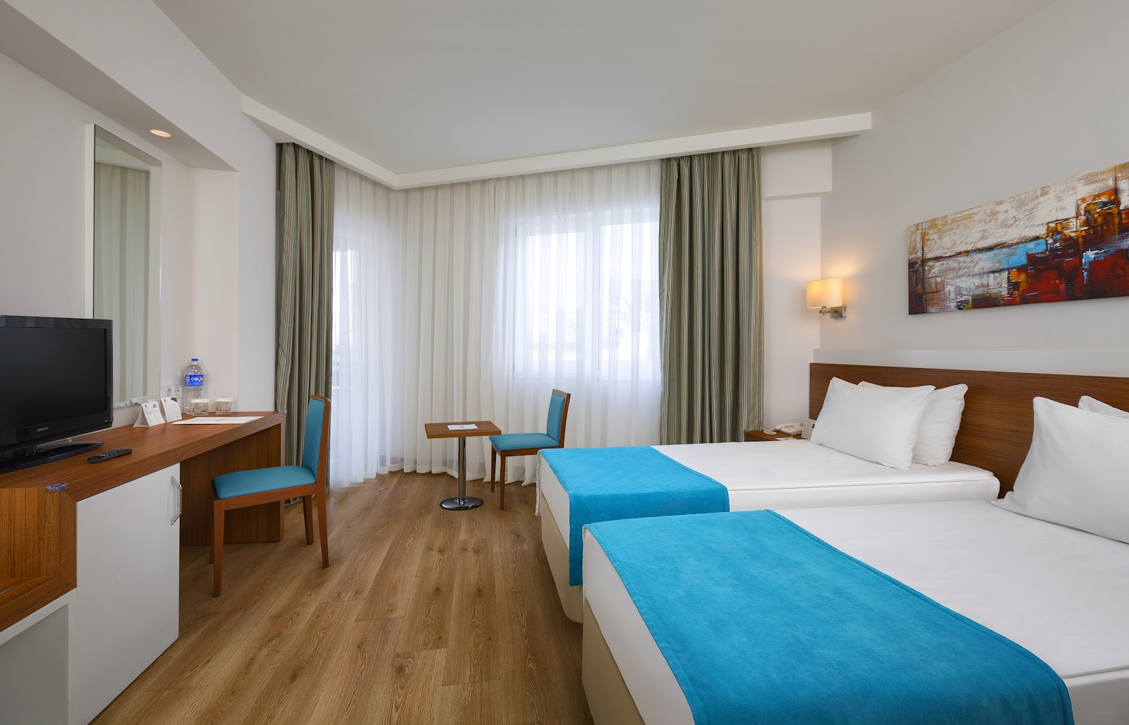 Resort hotel room with two beds topped with blue runners, wooden desk with TV, blue chairs, and floor-to-ceiling curtained windows.