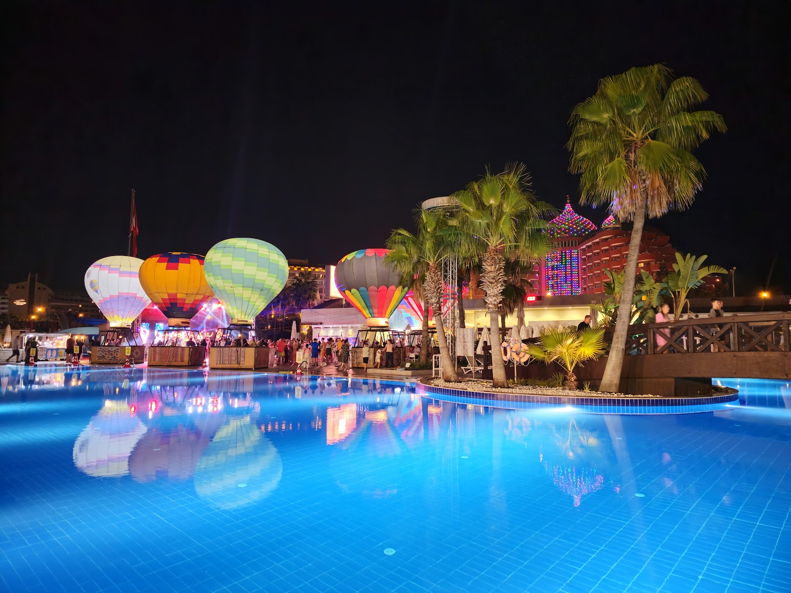 Illuminated hot air balloons float above a glowing blue resort pool at night, with palm trees and a colorful lit building visible in the background.