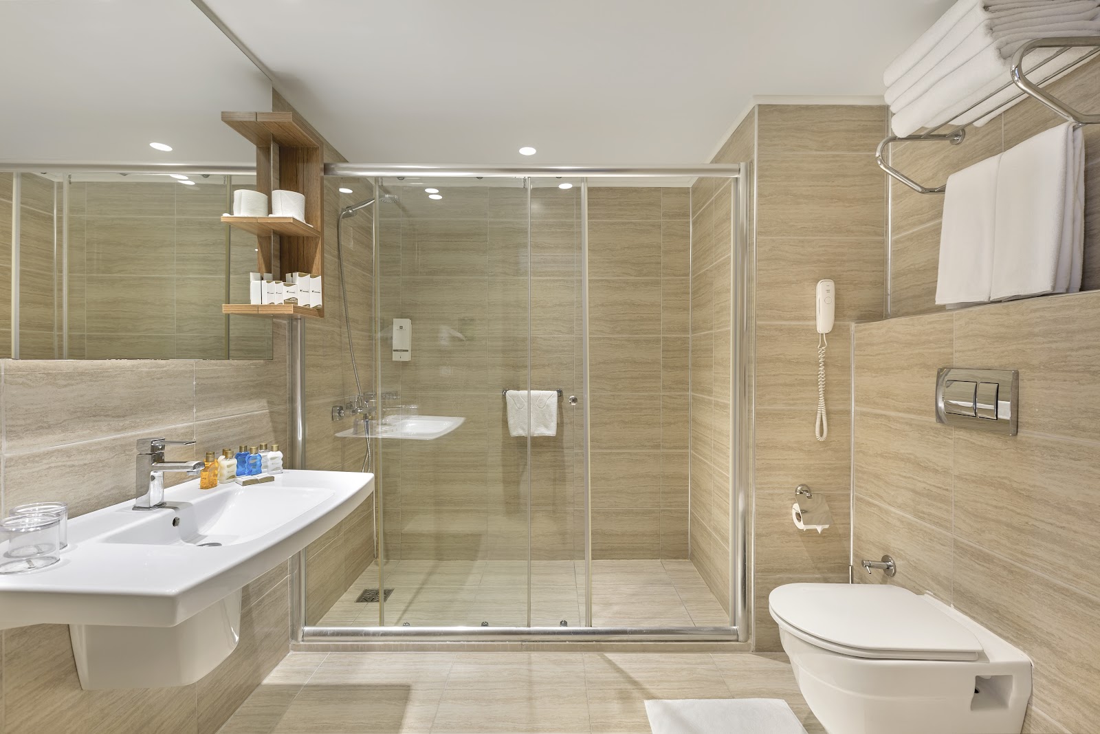 Modern bathroom with beige tile walls, glass-enclosed shower, white pedestal sink, and toilet at Corendon Grand Park Lara.