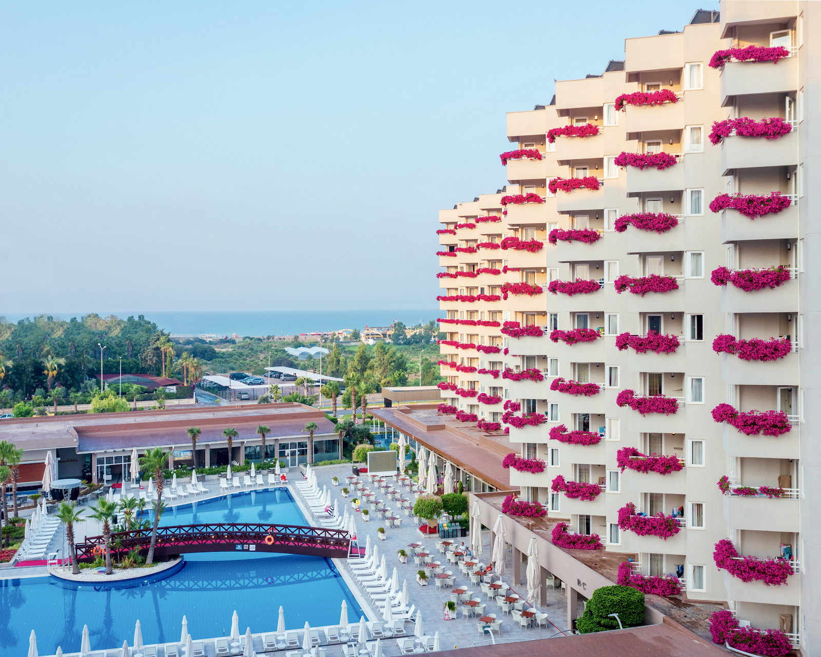 Cream-colored resort building with vibrant magenta bougainvillea cascading down balconies, overlooking a blue pool complex and Mediterranean coast.