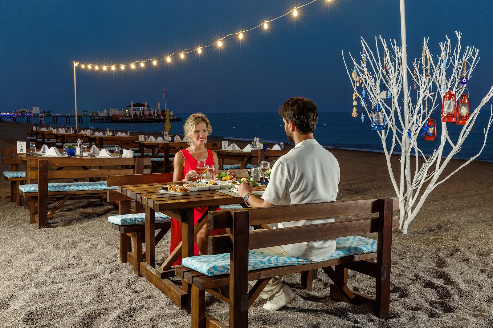 Couple dining at a beachfront table on sand at dusk, with string lights overhead, white decorative branches, and the sea beyond.