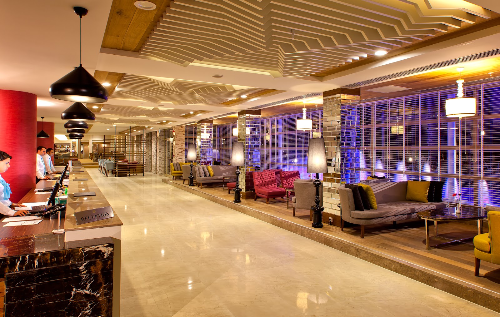 Modern lobby with polished marble floors, reception desk on left, seating areas with red and gray furniture, geometric wood ceiling, blue accent lighting, and large windows.
