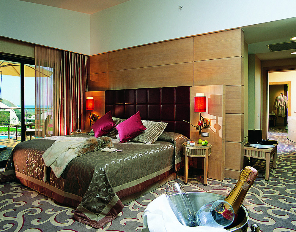 Modern resort bedroom with king bed, pink pillows, wood headboard, red lamps, ocean view balcony, and champagne bucket on patterned carpet.