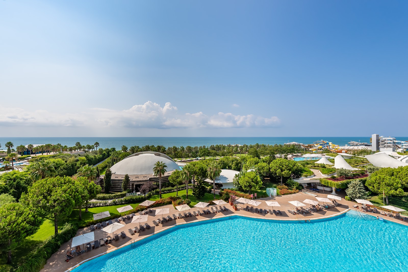 Turquoise pool with lounge chairs and umbrellas overlooks palm-lined grounds, dome structure, and Mediterranean Sea under clear sky.