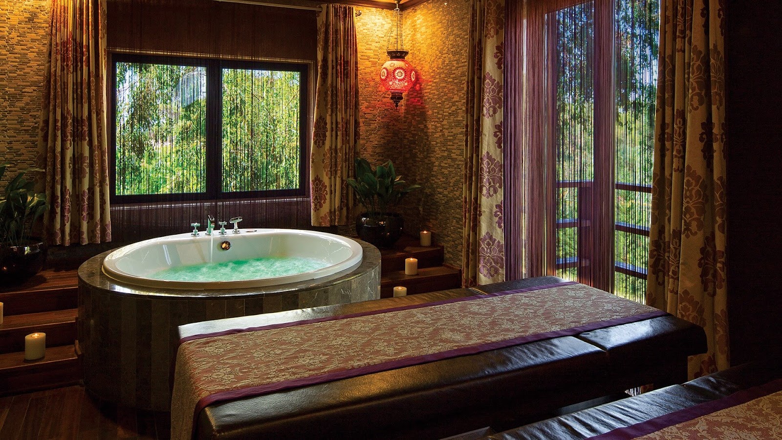 Spa bathroom with a round soaking tub filled with green water, massage bed in foreground, brick walls, red hanging lantern, and garden views through floor-to-ceiling windows.