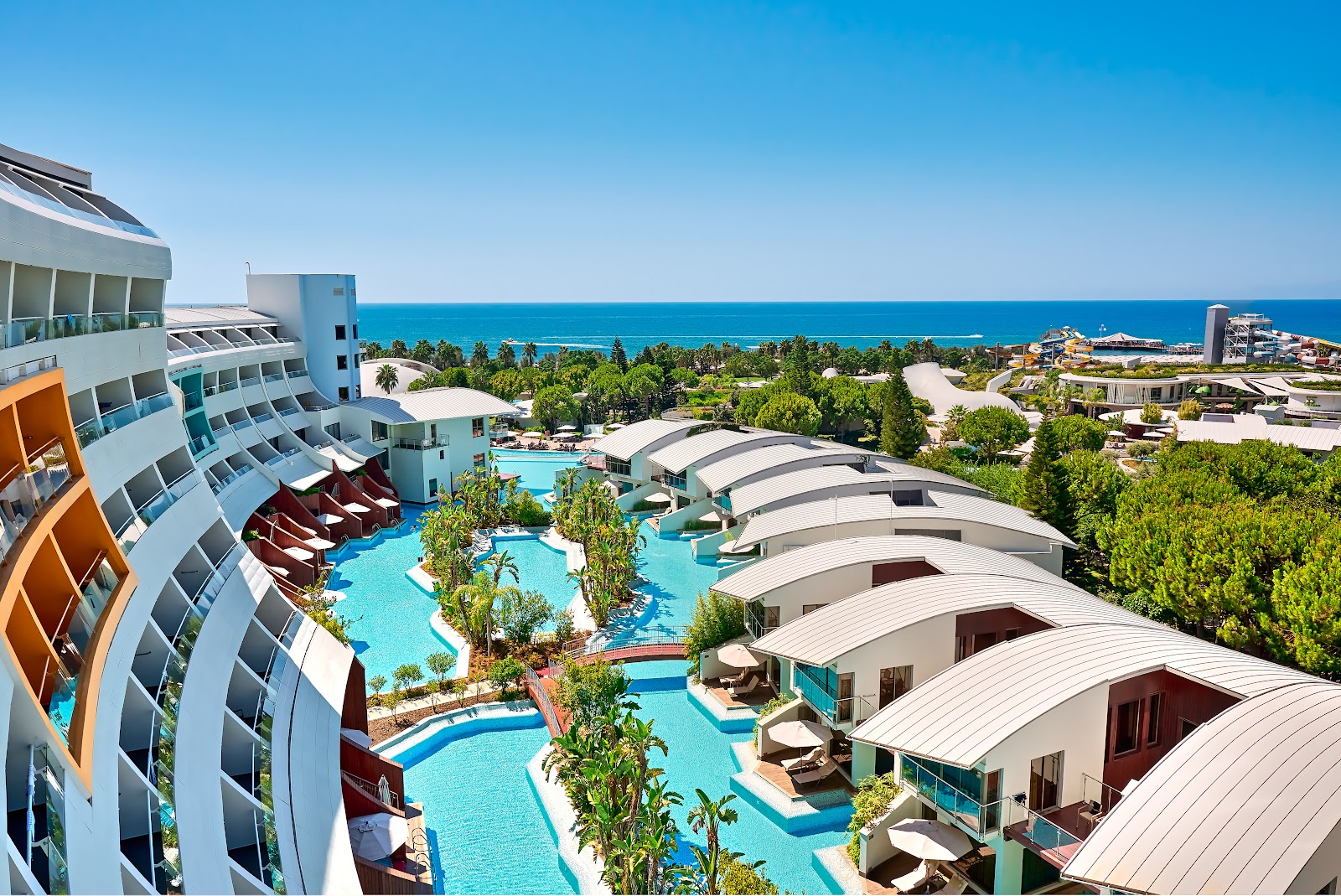 Aerial view of Cornelia Diamond Golf Resort & Spa showing curved hotel buildings, bright blue pools with loungers, palm trees, and the turquoise Mediterranean Sea beyond.