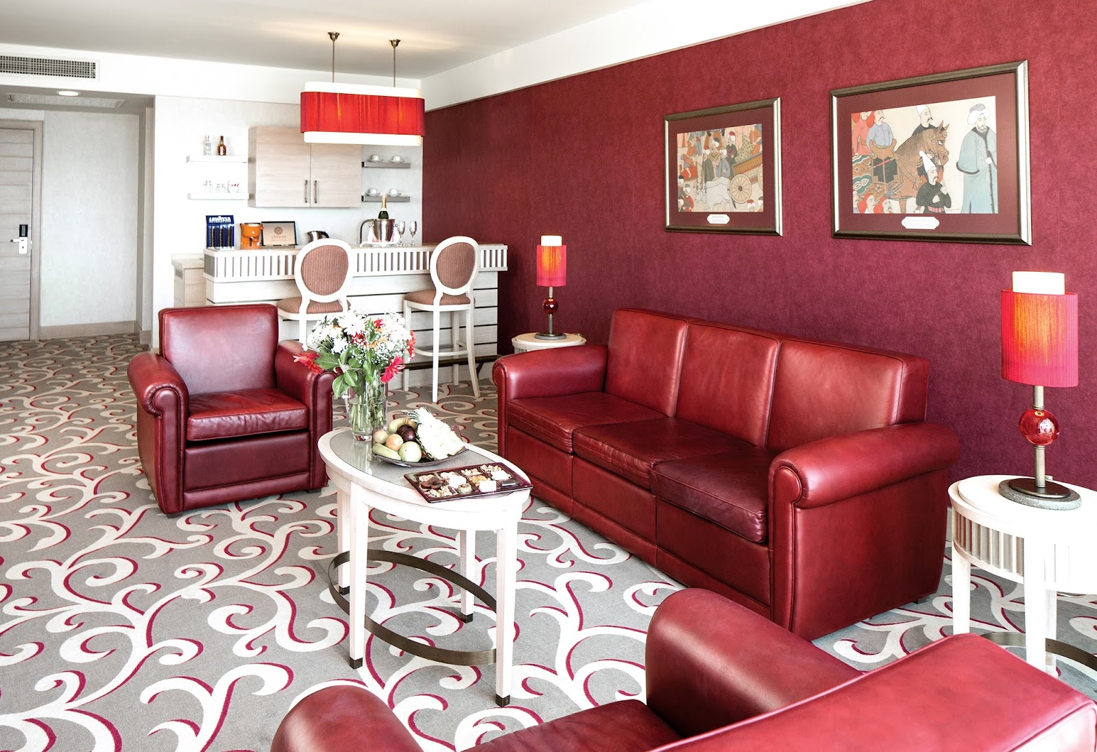A suite living area with burgundy walls, red leather furniture, patterned carpet, and a kitchenette with white cabinetry and red pendant lamp.