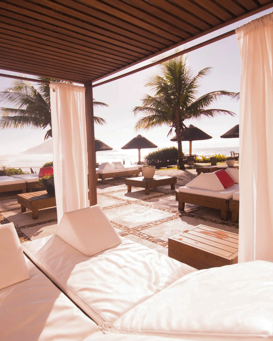 Covered lounge area with white cushioned daybeds overlooking palm trees, thatched umbrellas, and the beach at Costao do Santinho Resort.