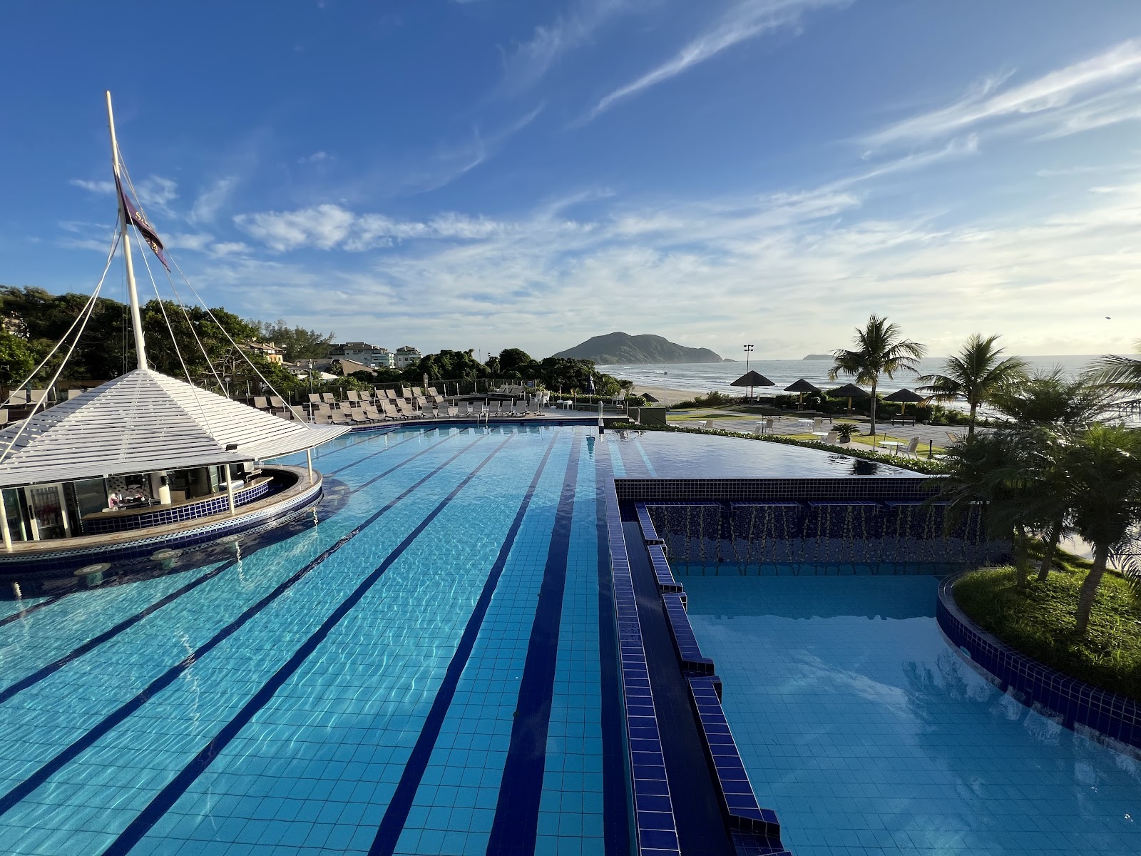 Lap pool with lane markers and adjoining leisure pool at Costao do Santinho Resort, with ocean and mountains visible beyond the palm-lined grounds.