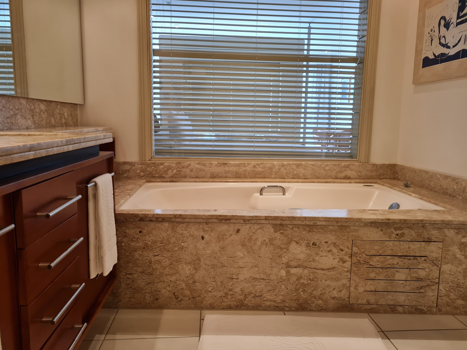 Bathroom with large soaking tub surrounded by travertine countertop, wood vanity, and window with shutters at Costao do Santinho Resort.