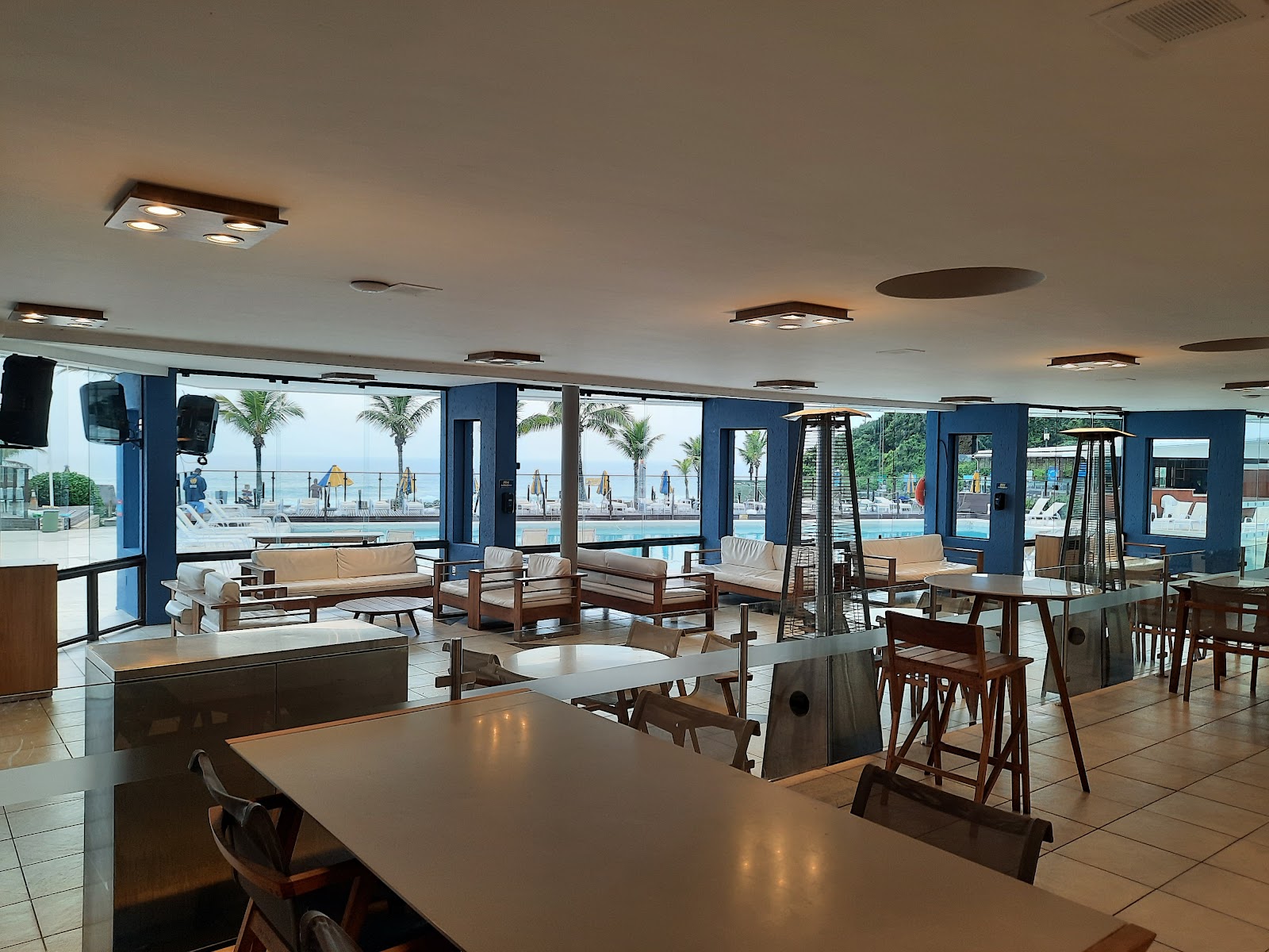 Indoor lounge and dining area with blue walls, wooden furniture, and floor-to-ceiling windows overlooking a beach with palm trees and ocean at Costao do Santinho Resort.