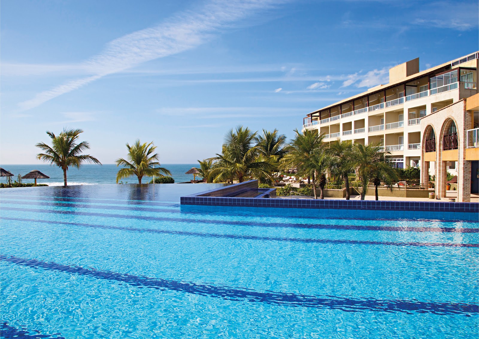 Infinity pool with swim lanes overlooking the ocean at Costao do Santinho Resort, flanked by palm trees and a multi-story building.