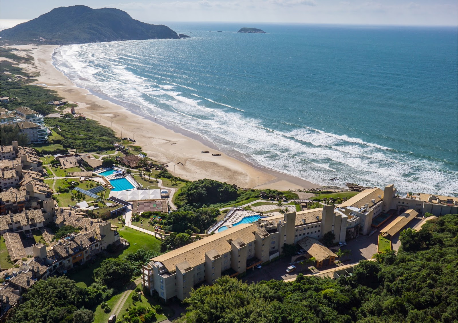 Aerial view of Costao do Santinho Resort with tan buildings, two pools, and a wide sandy beach with rolling waves backed by forested hills and distant rocky islands.