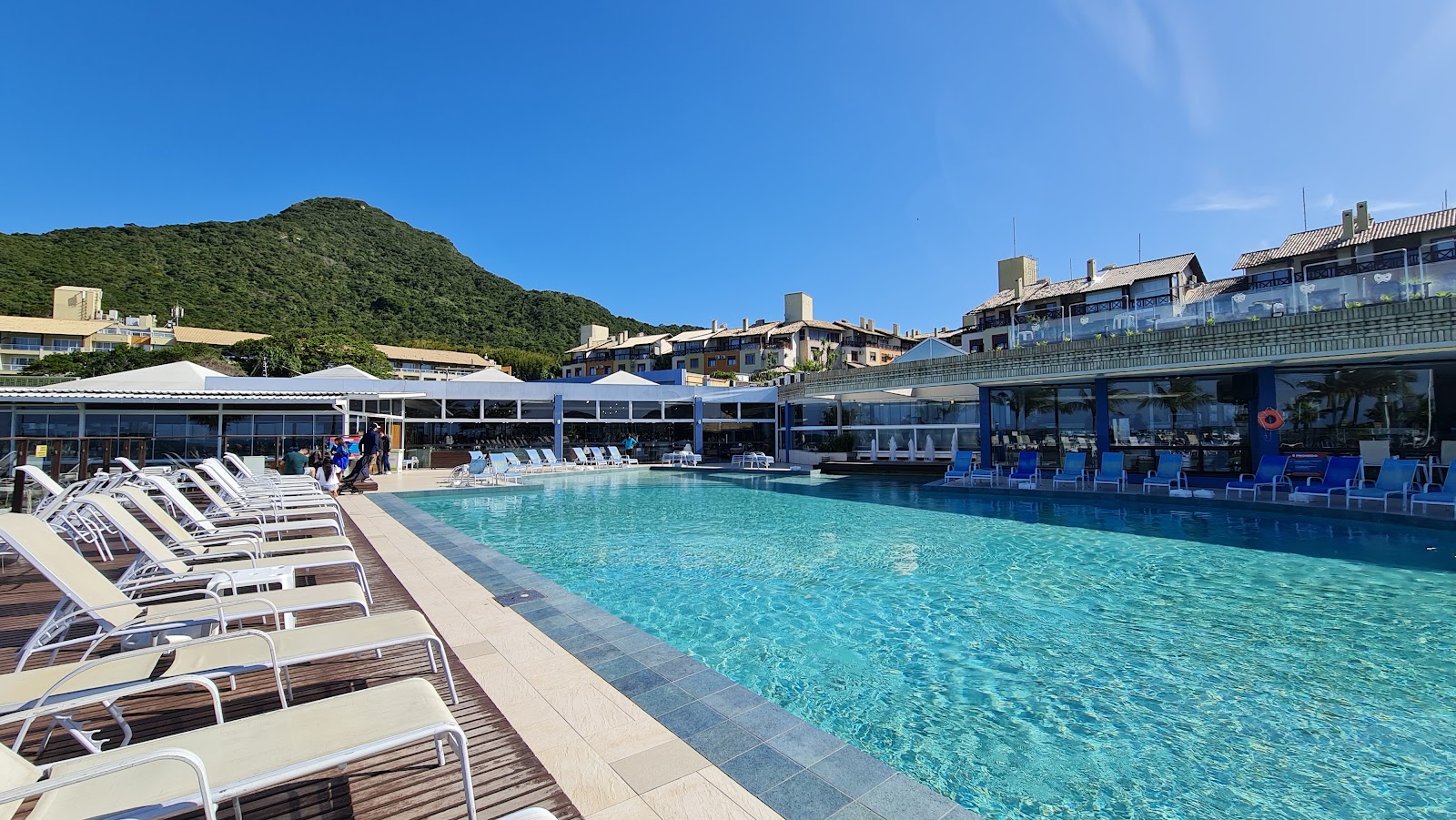 Rows of white lounge chairs line the pool deck at Costao do Santinho Resort, with a turquoise pool, covered bar area, and forested hillside beyond.