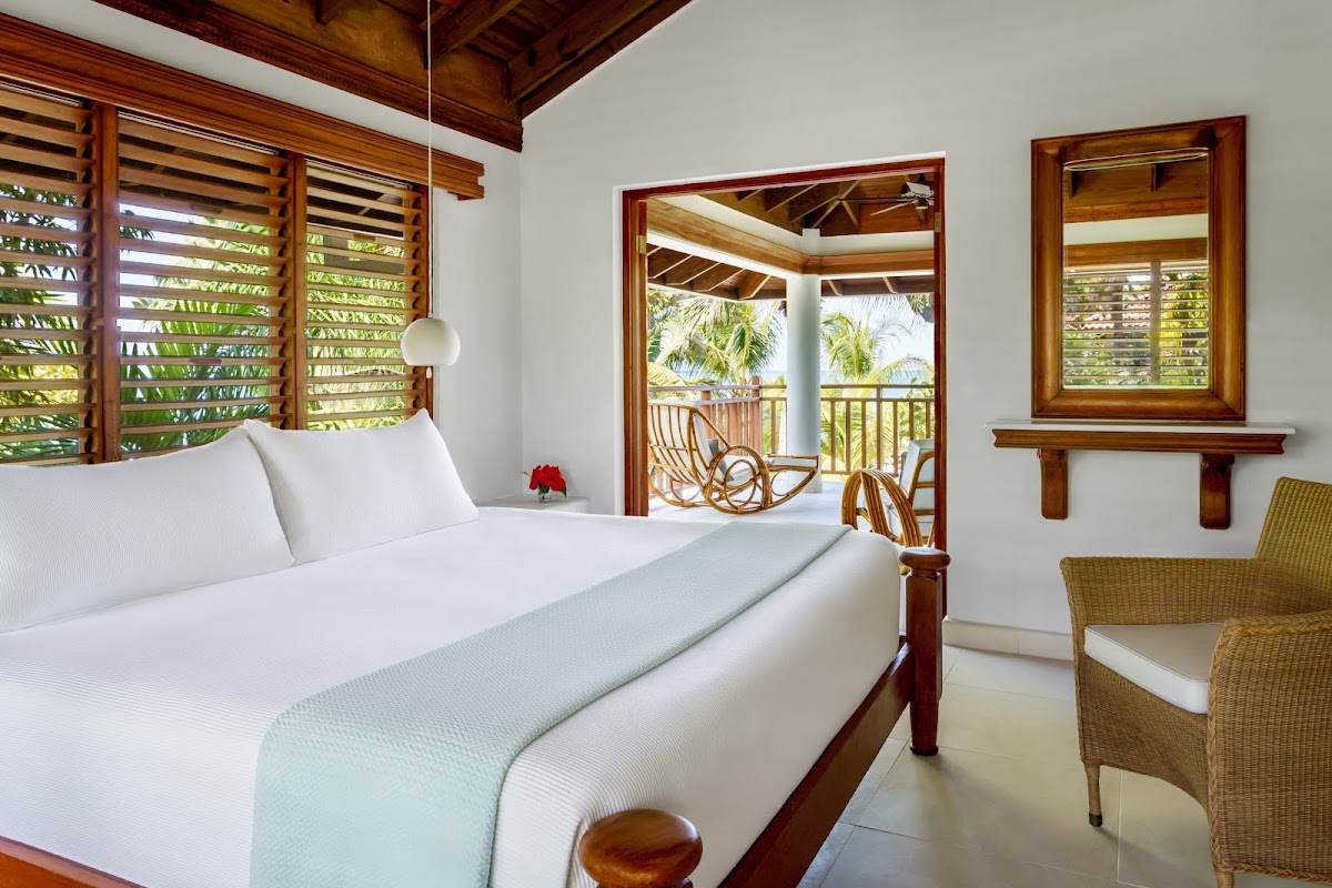 Bedroom with white linens on a wooden bed, wicker chair, louvered shutters, and a balcony overlooking palm trees.