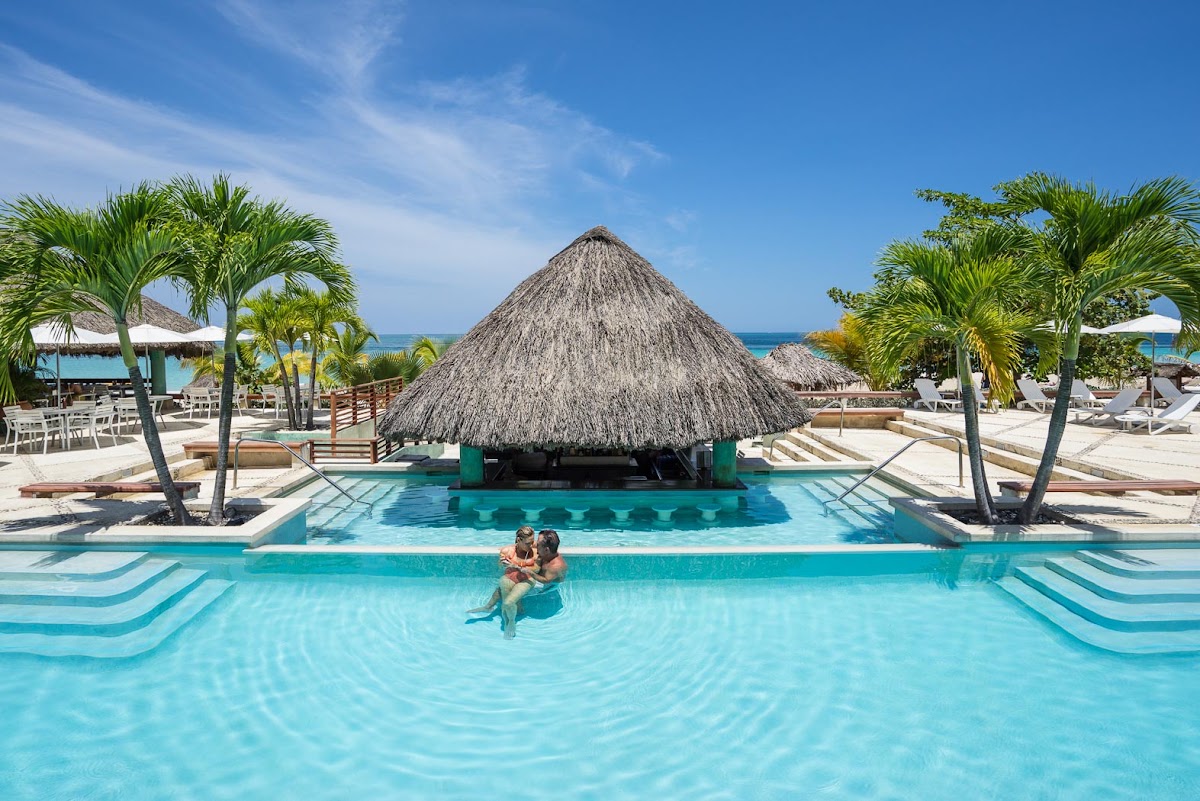 A couple floats in a turquoise pool leading to a large thatched-roof palapa, flanked by palm trees and loungers under the Caribbean sky.