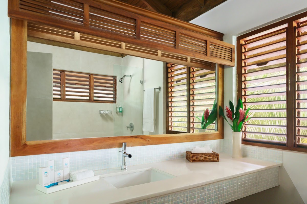 Bathroom with wooden-framed mirror, double sink vanity, glass shower enclosure, and wooden louvered windows overlooking tropical greenery.