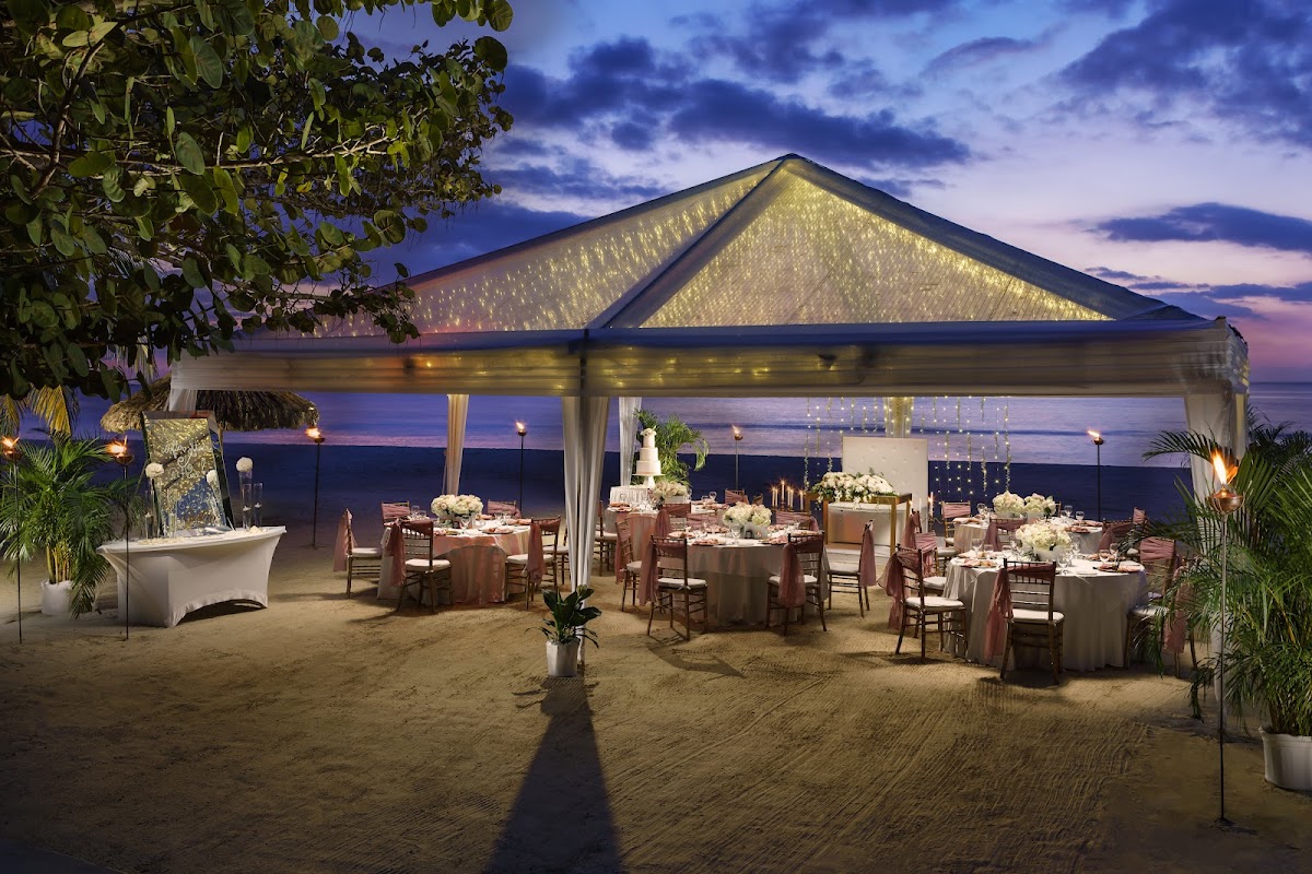 Illuminated beachfront pavilion with dining tables set for an event at dusk, ocean and twilight sky beyond.
