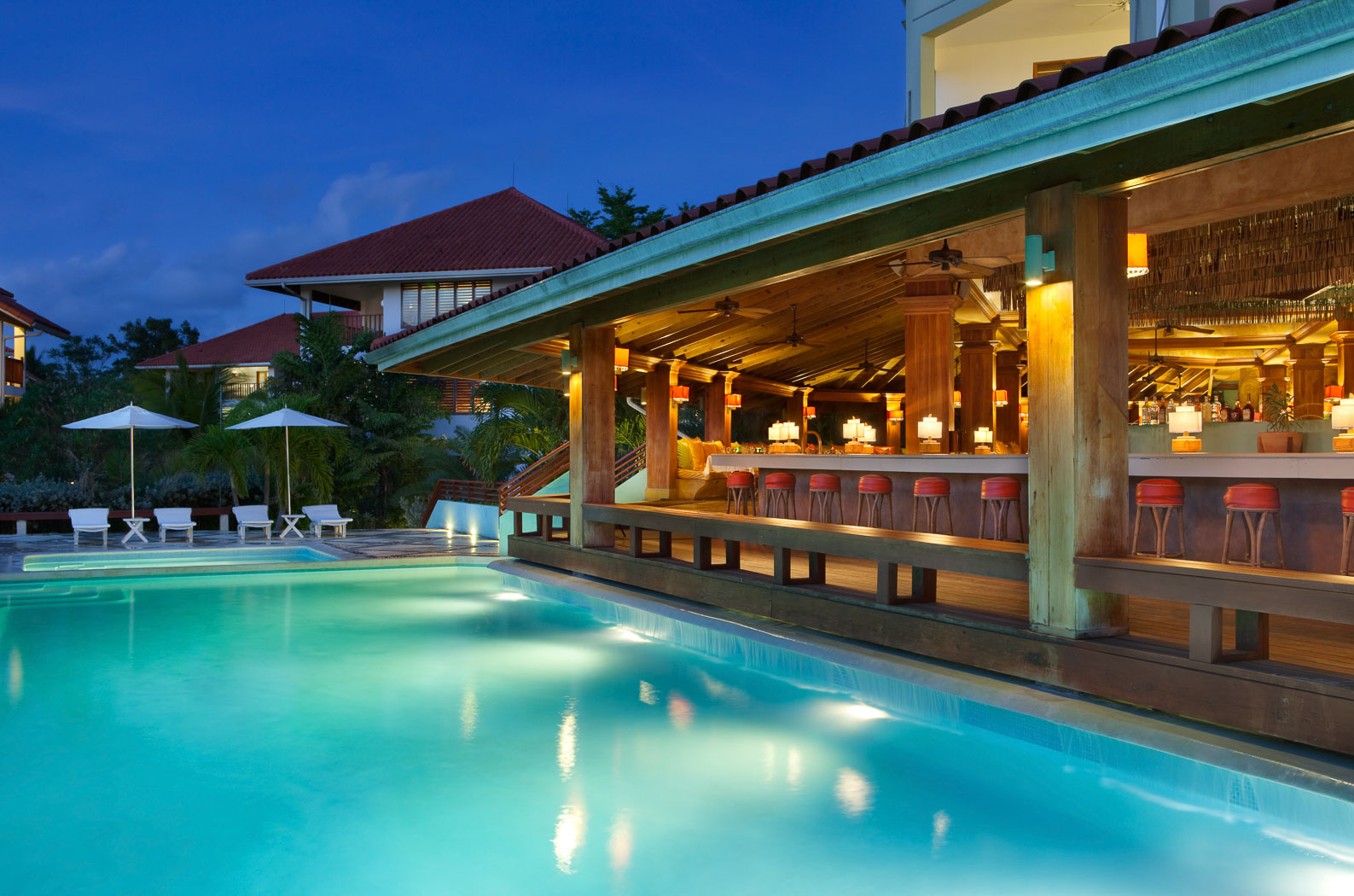 Illuminated poolside bar with red stools overlooks turquoise pool at dusk, with lounge chairs and umbrellas along the deck at Couples Swept Away.
