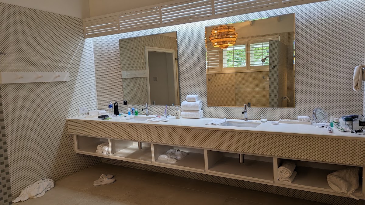 Resort bathroom with dual vanity, large mirror, woven pendant light, and open shelving below displaying rolled towels.