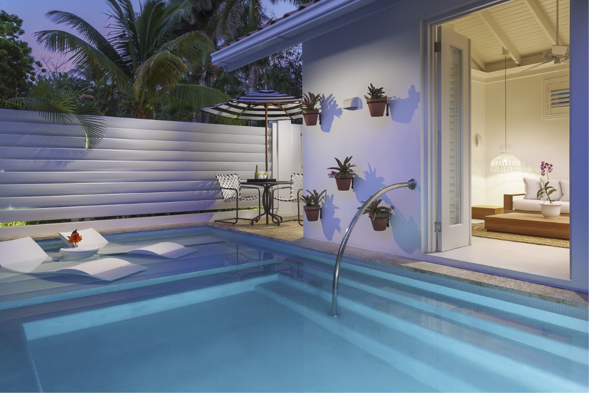 Private pool courtyard at dusk with loungers, umbrella, potted plants on white wall, and open suite door revealing bedroom interior.