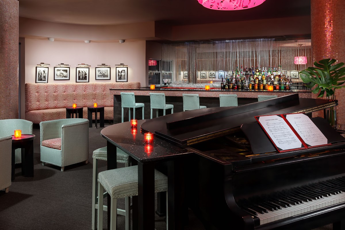 A grand piano with sheet music sits in a lounge area with mint-green chairs, pink neon lighting, a full bar, and framed photographs on brick walls.