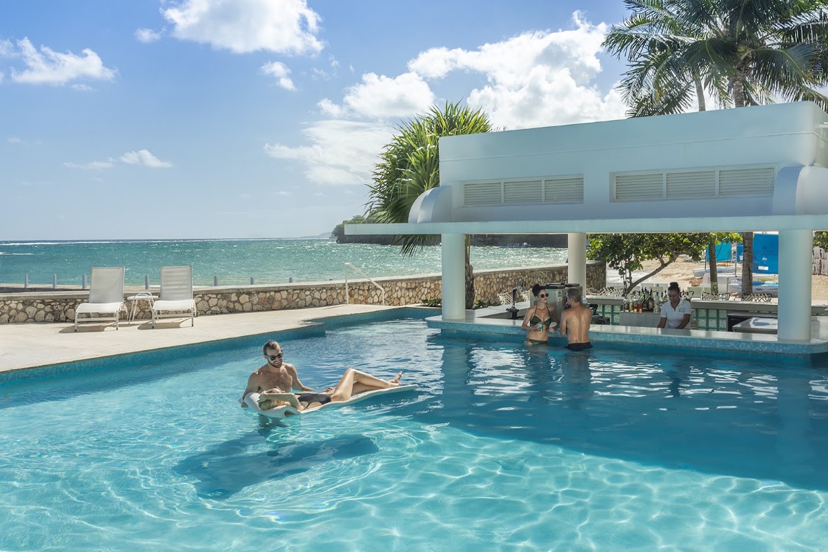Guests swim and relax in a beachfront pool at Couples Tower Isle, with the Caribbean Sea and palm trees visible beyond.
