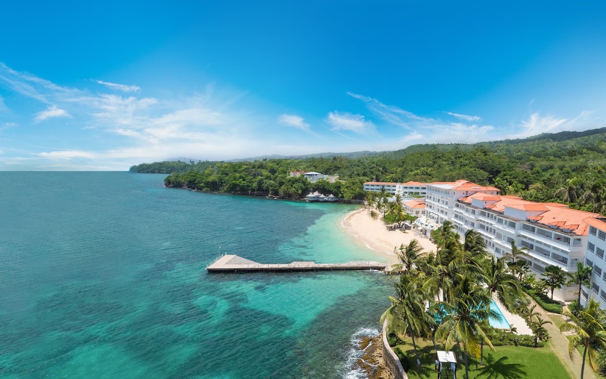 Aerial view of Couples Tower Isle resort with white multi-story buildings, private beach, turquoise water, and forested mountains under clear blue sky.