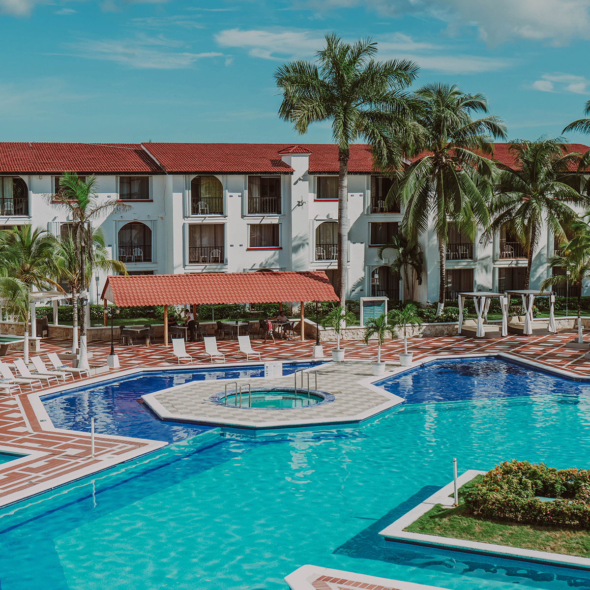 White building with red-tiled roof and palm trees overlooks turquoise swimming pools and hot tub surrounded by lounge chairs and patterned deck at Hotel Cozumel & Resort.