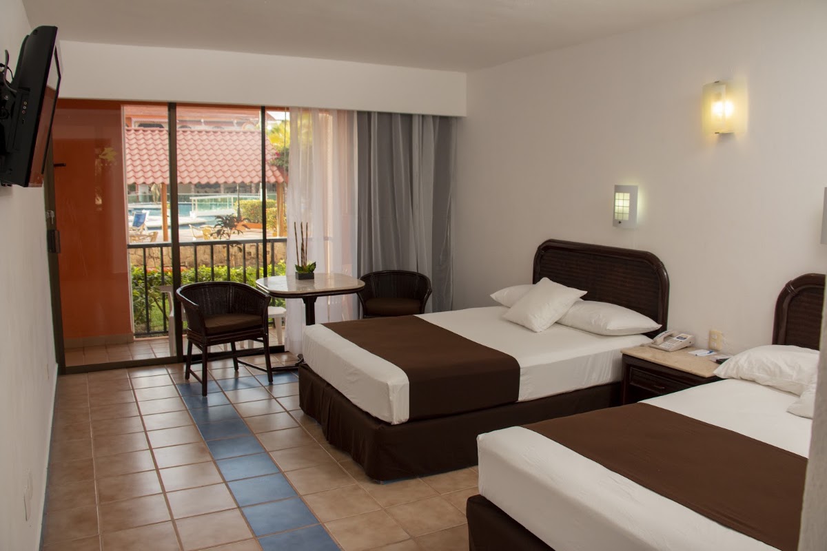 Hotel Cozumel & Resort guest room with two beds, seating area, and balcony overlooking pool and terracotta-roofed buildings.