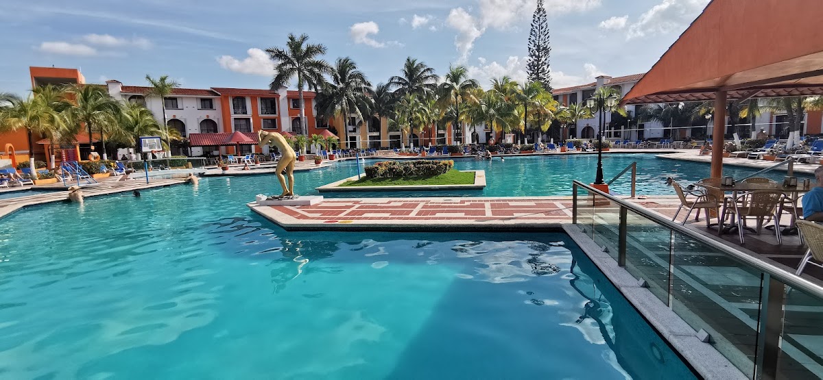 Turquoise pool with diving platform at Hotel Cozumel & Resort, surrounded by orange and white buildings, palm trees, and lounge chairs under pergolas.