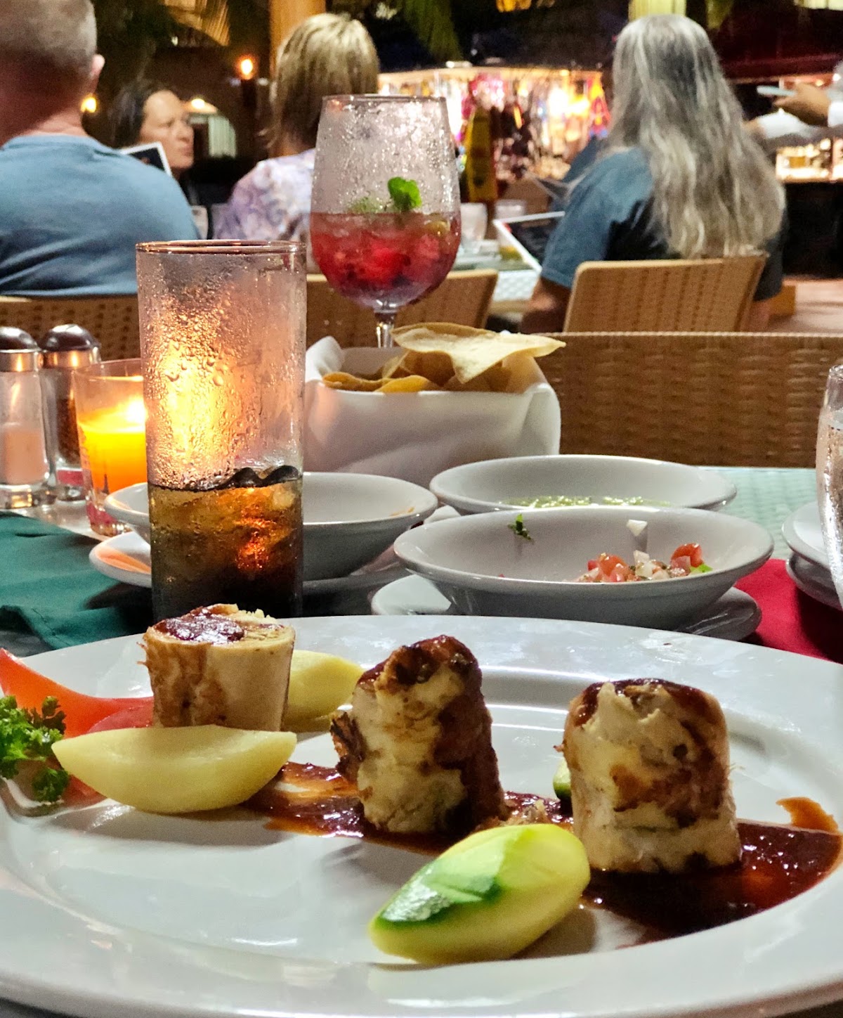 Plated seafood with seared scallops and avocado at an evening dining table, with cocktails and diners in the background.