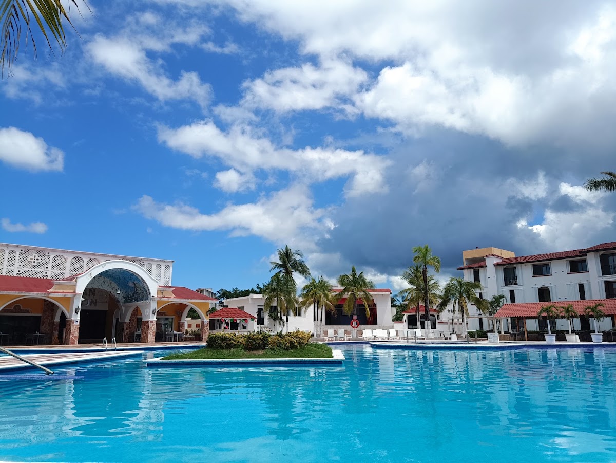 Turquoise pool with a floating platform, flanked by colonial-style buildings with red roofs and palm trees under blue sky at Hotel Cozumel & Resort.
